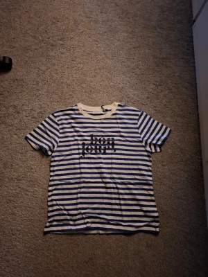 Randig t-shirt med tryck från Gina Tricot - Säljer en vit och blå randig t-shirt från Gina Tricot med texten 'bonjour!' tryckt i svart på bröstet. Klassisk rund halsringning och korta ärmar. Perfekt för dig som gillar fransk vibe och enkel stil.