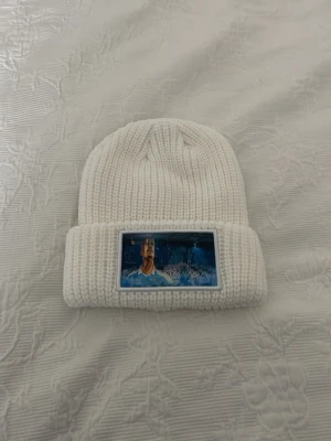 Vit stickad Malmö Ff Rosenberg mössa - Säljer en vit stickad mössa med ett färgstarkt tryck framtill som visar en person i blått. Mössan är ribbstickad och har en bred uppvikt kant. Perfekt accessoar för kyliga dagar.