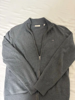 Grå kofta med dragkedja från GANT - Säljer en stilren grå kofta från GANT med hel dragkedja och ribbade muddar. Klassisk ståkrage och diskret GANT-logo broderad på bröstet. Perfekt till lager på lager och funkar året runt.