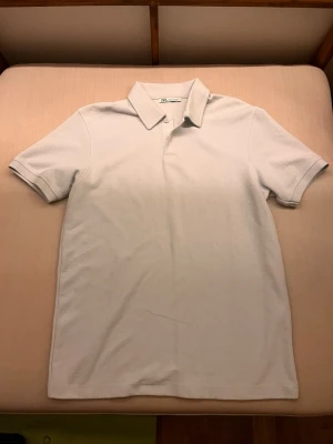 Ljusvit piké från Zara - Klassisk ljusvit pikétröja från Zara i storlek S. Skjortan har korta ärmar, krage och knappar framtill. Tillverkad i mjuk bomull som känns skön mot huden. Enkel och stilren design som passar till det mesta.
