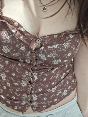 Brun blommig korsettopp med knappar - Supersöt brun korsettopp med vita blommor och träknappar hela vägen framtill. Axelbanden är volangiga och toppen har en figurnära passform. Perfekt för dig som vill ha en romantisk och trendig look i sommar. 