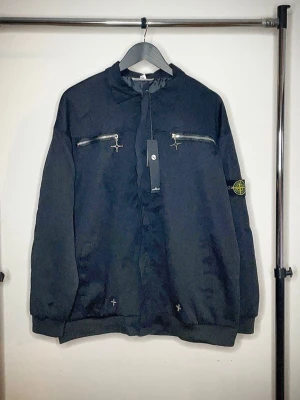 Svart bomberjacka från Stone Island - Svart bomberjacka från Stone Island med flera silvriga dragkedjor på bröstet, ärmen och ryggen. Jackan har klassisk krage, ribbade muddar och detaljer med vita korsstygn vid nederkanten. Materialet är polyester och jackan har en loose passform.