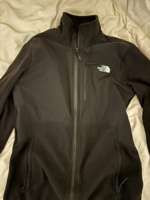 Svart fleecejacka från The North Face - Säljer en svart fleecejacka från The North Face med hög krage och dragkedja framtill. Jackan har en bröstficka med dragkedja och två sidofickor. Perfekt för kyliga dagar och enkel att matcha med allt.