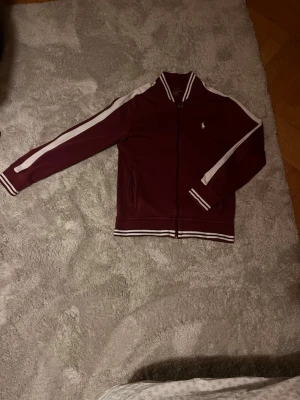 Vinröd zip hoodie från Polo Ralph Lauren - Säljer en vinröd hoodie från Polo Ralph Lauren med vita ränder längs ärmarna och på kragen. Tröjan har dragkedja framtill, två fickor och ribbade muddar. Minimala skrapor på sina ställen (defekt vid handleden) men ingenting som märks. 