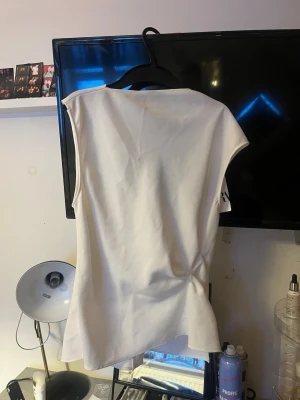 Vit blus med knappar från Zara - Säljer en vit blus från Zara med coola axelvinklar och knappar längs hela ryggen. Blusen är ärmlös och har en öppen ryggdetalj som ger en unik look. Perfekt till svarta jeans eller kjol för en clean och trendig stil. Köpt på plick men tyvärr fel storlek