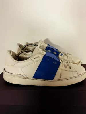 Valentino Open sneakers vit/blå - Säljer ett par Valentino Open sneakers i vitt skinn med bred blå rem över mitten. Klassisk låg modell med rund tå, snörning och subtila nitar på hälen. Perfekta för dig som vill ha en clean men ändå unik look. Medföljs box+ dustbag 