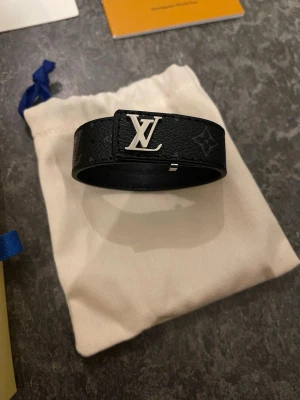  svart monogram läderarmband - Säljer ett svart armband i präglat läder med klassiskt monogrammönster och silverfärgat spänne. Snygg accessoar som lyfter vilken outfit som helst. Kommer med original ask och dustbag.