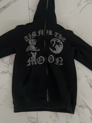 Rhinestones hoodie ”aim for the moon” - Rhinestones hoodie från Aimforthemoon med dragkedja och strasstenar som bildar motiv av en beväpnad figur och en jordglob med texten ”Aim for the moon”. Storlek: m
