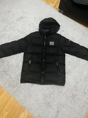 Svart pufferjacka från EA7 Emporio Armani - Säljer en svart pufferjacka från EA7 Emporio Armani med huva och dragkedja framtill. Jackan har två fickor med dragkedja och tydlig logga på bröstet. Perfekt för kalla dagar och riktigt snygg streetstil.
