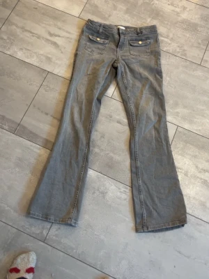 Grå bootcut jeans med fickdetaljer - Säljer ett par gråa jeans med bootcut passform och snygga fickor framtill med knappdetaljer. Klassisk femficksmodell och bälteshällor. Perfekta för dig som gillar en lite vidare siluett nertill. Materialet är jeans och färgen är en neutral grå.