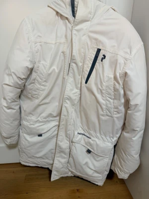 Vit Peak Performance vinterjacka - Säljer en vit pufferjacka från Peak Performance med huva och fuskpäls. Jackan har flera fickor med dragkedja och Peak Performance-logga på bröstet och ärmen. Perfekt för kalla dagar med sin tjocka vaddering och snygga detaljer.