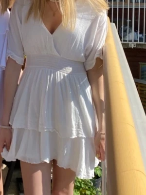 Vit omlottklänning från Amisu - Supersöt vit kortklänning från Amisu med omlott-v-ringning, smock i midjan och volangdetaljer. Klänningen har korta ärmar med lätt puff och är gjord i ett tunt, luftigt material som känns perfekt för varma dagar.