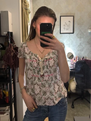 Mönstrad blus med spetsdetaljer - Supersöt blus med korta puffärmar och v-ringning. Blusen har ett unikt mönster i ljusrosa, beige och grönt med små spetsdetaljer och knappar framtill🥰