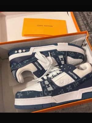 Louis Vuitton blå/vita sneakers - Säljer ett par Louis Vuitton sneakers i blått och vitt med klassiskt monogrammönster. Skorna har snörning, platt sula och detaljer i både skinn och textil. Snygg kontrast mellan marinblå och vita partier samt tydlig LV-logga på sidan och sulan.