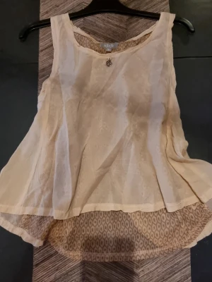 Gammelrosa ärmlös linnetopp från ajlajk - Säljer en gammelrosa ärmlös linnetopp från ajlajkajit med rund halsringning och ett dekorativt silverfärgat hänge framtill. Toppen har ett lätt transparent yttertyg och ett mönstrat innerfoder i beige toner. Baktill finns en öppen detalj som ger en luftig känsla.