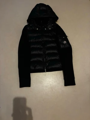 Svart Moncler pufferjacka med huva - Säljer en svart pufferjacka från Moncler med huva och dragkedja framtill. Jackan har quiltad kropp i blankt material och stickade ärmar. På ena ärmen finns en ficka med Moncler-logga. Perfekt för dig som vill ha en clean och stilren look.