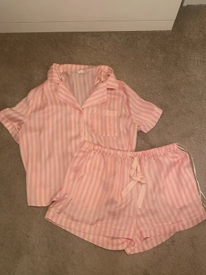 Rosa och vita randiga pyjamas från Primark - Supersöt pyjamas från Primark i ljusrosa och vitt med ränder. Setet består av en kortärmad skjorta med bröstficka och matchande shorts med dragsko i midjan. Materialet är mjukt och skönt, perfekt för chillkvällar eller mysiga morgnar.