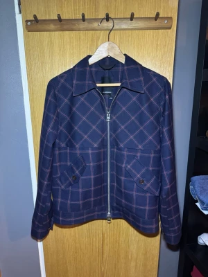 Rutig blå overshirt från J.Lindeberg - Snygg overshirt från J.Lindeberg i mörkblått med rött rutmönster. Jackan har klassisk krage, två fickor med knapp framtill. Materialet är grovvävt och känns robust, perfekt för lager på lager. Passar dig som gillar stilrena detaljer.