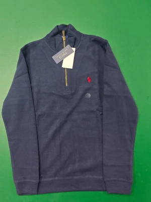 Marinblå half zip tröja från Polo Ralph Lauren - Snygg marinblå långärmad tröja från Polo Ralph Lauren med guldfärgad half zip-dragkedja och röd broderad logga på bröstet. Tröjan har en klassisk passform och är gjord i mjuk bomull, perfekt för lager på lager under höst och vinter.