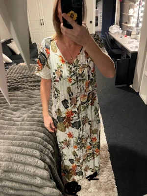 Blommig långklänning från Emery Rose - Säljer en superfin långklänning från Emery Rose i storlek S. Klänningen har ett blommigt mönster i grönt, rött, beige och svart på en vit botten. Den har korta ärmar, v-ringad hals och är gjord i ett lätt och luftigt material, perfekt för varma dagar.