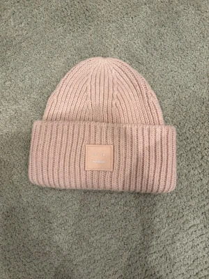 Ljusrosa mössa från Acne Studios - Säljer en ribbstickad ljusrosa mössa från Acne Studios med det klassiska face-loggan framtill. Mössan är i mjuk ull och har en bred uppvikt kant. Perfekt accessoar för att hålla värmen och addera lite stil till din outfit.