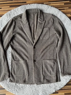 Brun kavaj/stickad cardigan från John Henric - Snygg brun cardigan från John Henric i 100% bomull. Cardiganen har två knappar framtill, breda slag och tre praktiska fickor. Materialet är mjukt och har en diskret struktur som ger en chill och modern vibe.