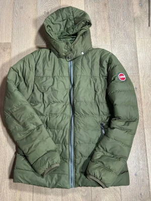 Grön pufferjacka från Colmar - Säljer en grön pufferjacka från Colmar med huva och dragkedja framtill. Jackan har quiltad design, långa ärmar och två fickor med dragkedja. Perfekt för kalla dagar och riktigt skön att bära. Klassisk logga på ärmen.
