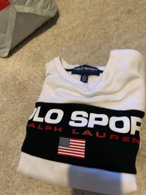 Vit och svart sweatshirt Polo Sport - Säljer en clean vit sweatshirt från Polo Sport Ralph Lauren med svart band över bröstet och armarna. Stor logga framtill och amerikansk flagga som detalj. Tröjan har rund hals och långa ärmar, perfekt för en sportig och avslappnad look.