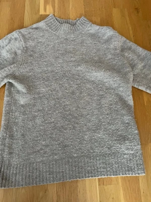 Beige stickad tröja Pull&Bear - Mysig grå stickad tröja från Pull&Bear med hög ribbad krage och långa ärmar. Tröjan har en relaxed passform och ribbade muddar vid ärmslut och nederkant. Perfekt för kyliga dagar och enkel att styla till jeans eller kjol.