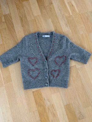 Grå kofta med hjärtan från Zara - Superfin grå kofta från Zara med röda pärlbroderade hjärtan framtill och baktill. Koftan har v-ringning, knappar framtill och är dekorerad med små pärlor i vitt och rött. Mjuk och stickad, perfekt för kyliga dagar. Använd en gång 