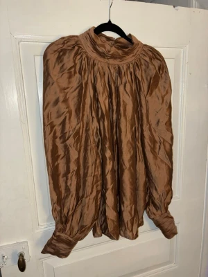 Brun satinblus H&M - Elegant brun blus i satinliknande material med hög hals, rynkad front och voluminösa ärmar med dekorativa manschettknappar. Från H&M, strl L.