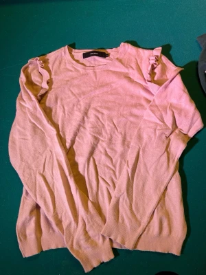 Ljusrosa tröja med volang Vero Moda - Supersöt ljusrosa tröja från Vero Moda i storlek XL. Tröjan har långa ärmar och volangdetaljer på axlarna som ger en extra touch. Mjuk och skön, perfekt till både jeans och kjol. Rund halsringning och enkel, clean look.