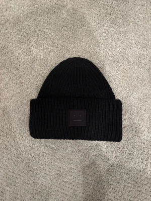 Svart mössa från Acne Studios - Säljer en svart ribbstickad mössa från Acne Studios med det klassiska ansiktslogotypen framtill. Mössan är i tjockt, mjukt material och har en bred uppvikt kant. Perfekt för kalla dagar och ger en clean look.