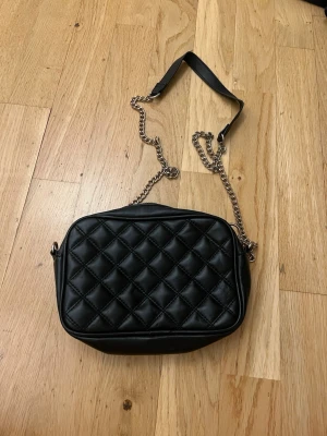 Svart quiltad axelväska med kedja från hm - Säljer en svart axelväska i skinnimitation med quiltat mönster. Väskan har en guldfärgad kedjerem med en del i svart material för axeln. Stängs med dragkedja och har en stilren, tidlös look som passar till många outfits. Har en liten puder fläck i.