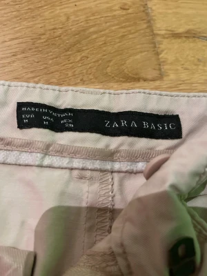 Beiga chinos från Zara Basic - Snygga beiga chinos från Zara Basic i storlek M. Byxorna har klassisk passform med hällor för bälte och diskreta veck framtill. Perfekta för en clean och stilren look. Tillverkade i mjukt bomullsmaterial som känns skönt mot huden.