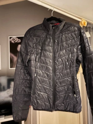 Svart pufferjacka - Säljer en svart pufferjacka med quiltad design och dragkedja framtill. Jackan har hög krage, två fickor med dragkedja och en diskret rosa detalj på insidan. Perfekt för kyliga dagar och enkel att matcha med olika outfits. Jackan har aldrig används, den har bara legat i garderoben ett tag.