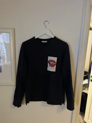 Svart sweatshirt Limitato x Sara Pope - Cool mörk blå sweatshirt från Limitato i samarbete med Sara Pope. Tröjan har ett unikt tryck med röda läppar på bröstet och är långärmad med rund hals. Materialet är mjukt och känns som bomull.