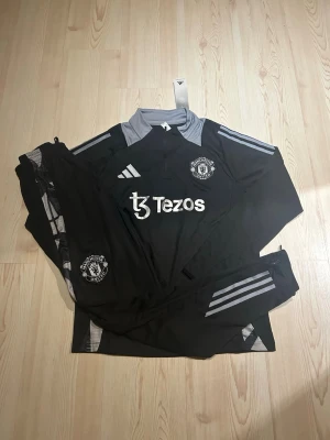 svart Manchester United - Storlek: S - Bilderna visar ett svart Manchester United träningsset från Adidas med grå detaljer och klubbmärke, Storlek : S