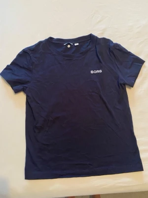 Mörkblå t-shirt från Björn Borg - En stilren mörkblå t-shirt från Björn Borg med enkel logga på bröstet. Klassisk passform och rund halsringning, perfekt till jeans eller shorts. Gjord i mjuk bomull som känns skön mot huden.