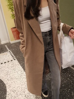 Beige lång kappa med bälte - Säljer en snygg beige kappa i lång modell med bälte i midjan. Jackan har breda slag och är helfärgad utan mönster. Perfekt för dig som vill ha en stilren och klassisk look. Passar till både jeans och sneakers. Original pris på bikbok 999 kr använd endast en gång 