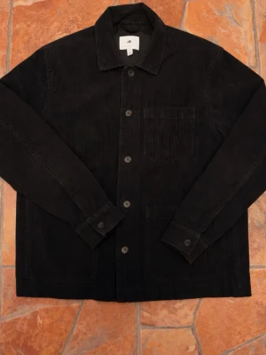 Svart manchester overshirt från H&M - Säljer en svart overshirt från H&M i mjuk manchester. Jackan har klassisk krage, knäppning framtill med fyra knappar och två stora fickor på framsidan. Perfekt lager-på-lager-plagg med lång ärm och enkel, stilren look.