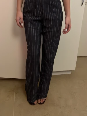 Svarta randiga kostymbyxor - Säljer ett par svarta kostymbyxor med vita tunna ränder. Byxorna har hög midja och ett rakt, lite vidare snitt som ger en chill och trendig look. Perfekta att styla med sneakers eller klackar. Materialet känns lätt och bekvämt.