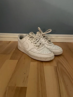 Nike court vision vita sneakers - Klassiska Nike court visionsneakers, som kan kopplas till tidslösa air force 1 i helvitt skinn med perforerad tå, rund tå och snörning. Lågt skaft och platt sula. Perfekta för dig som gillar en clean och tidlös look. Passar till det mesta och är riktigt bekväma.