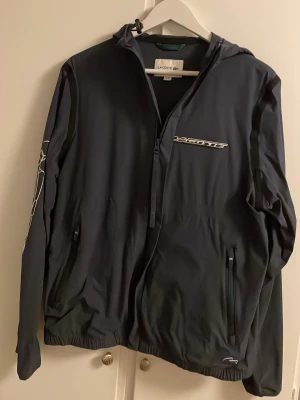 Svart vindjacka från Lacoste - Säljer en svart vindjacka från Lacoste med huva och dragkedja. Jackan har stora vita Lacoste-prints på bröstet och ena ärmen samt två fickor med dragkedja. Materialet är lätt och syntetiskt, perfekt för blåsiga dagar. Elastiska muddar vid ärmslut och nederkant. 9/10. Köpt för 2700kr i Lacoste egna butik.