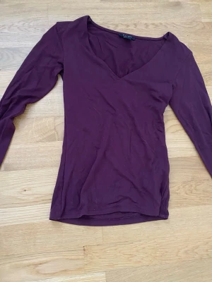 Vinröd långärmad topp från Amisu - Säljer en enkel och snygg vinröd långärmad topp från Amisu i storlek XS. Toppen har en djup v-ringning och är gjord i mjukt bomullsmaterial. Perfekt att styla med jeans eller kjol för en clean look.