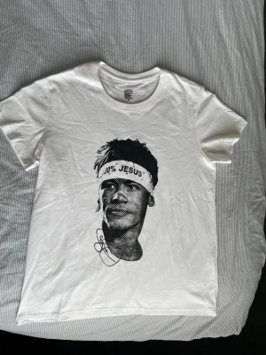 Vit t-shirt med svart tryck XS - Vit t-shirt i 100% bomull med svart grafiskt tryck av ett ansikte på framsidan och en stor svart sköld med pilar och stjärnor på ryggen. Klassisk rund hals och korta ärmar. Snygg och stilren design som sticker ut.