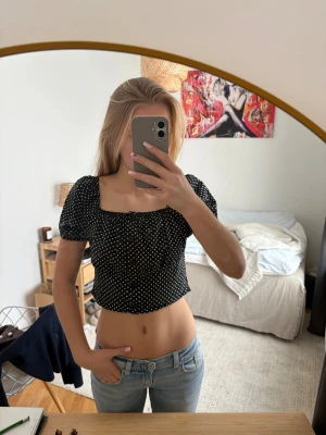Svart croppad prickig blus - Säljer en svart croppad blus med vita prickar och puffiga korta ärmar. Toppen har knappar framtill och smockad rygg för extra komfort. Den har en fyrkantig urringning och är gjord i ett lätt bomullstyg. Perfekt för en trendig och avslappnad look.❤️pris går att diskutera såklart!!