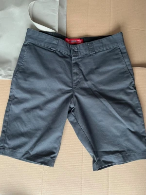 Gråa Dickies shorts  - Klassiska svarta shorts från Dickies med slim passform och raka ben. Tillverkade i slitstark bomull, har bälteshällor, knapp och dragkedja framtill samt två bakfickor. Perfekta för en clean och enkel stil.