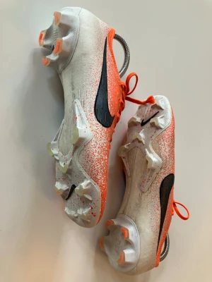 Nike Mercurial Elite  - Säljer ett par riktigt snygga Nike fotbollsskor i orange och vitt med svart swoosh på sidan. Skorna har snörning, platt sula med dobbar och coolt stänkmönster över hela ovandelen. Perfekta för dig som vill sticka ut på planen!