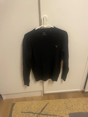 Svart kabelstickad tröja från Gant - Svart kabelstickad tröja från Gant i premium bomull. Klassisk rund halsringning och diskret Gant-logga broderad på bröstet. Långärmad modell med ribbade muddar och en riktigt clean look. Perfekt för dig som gillar stilrena vibes.